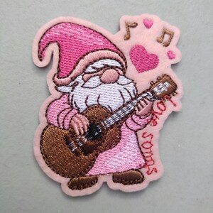 May include: A pink and brown embroidered patch featuring a gnome playing a guitar. The gnome wears a pink hat and outfit, with a white beard. The patch includes musical notes, a heart, and the words "love songs".