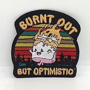 May include: A black and yellow embroidered patch with a retro sunset design. The patch features a cartoon character with a happy face and the text "Burnt Out But Optimistic".