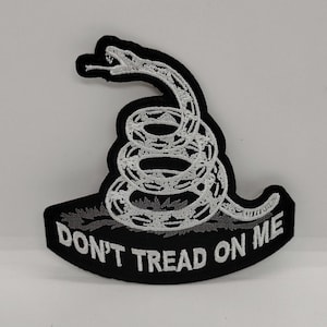 May include: A black and white embroidered patch featuring a coiled rattlesnake design. The snake is depicted in white thread with black detailing, positioned above a curved black banner containing the white text DON'T TREAD ON ME.