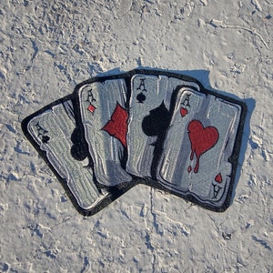 May include: Embroidered patch featuring four playing cards, each an Ace. The suits are diamond, spade, club, and heart. The heart suit has a dripping blood effect. The cards are silver with black borders.