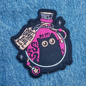 May include: Embroidered patch featuring a black cat inside a pink potion bottle. The patch includes the text "DO IT!" and "DON'T OPEN EVIL Inside". The patch has a black border and is set against a blue denim background.