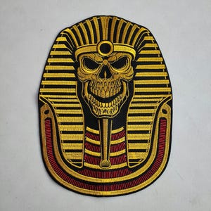 May include: Embroidered patch featuring a skull in a pharaoh headdress. The design uses gold, black, and red thread. The skull has a wide grin, and the headdress is detailed with vertical stripes. The patch is likely for clothing or accessories.