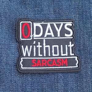 May include: A black and white embroidered patch with the text "0 DAYS without SARCASM" in white and red lettering. The patch is rectangular with a decorative border and is set against a blue denim background.