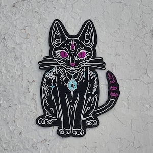 May include: Black embroidered patch of a cat with pink eyes and a turquoise gem on its chest. The cat has a detailed design with white lines and a pink tail.