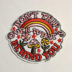 May include: Embroidered patch with the text "DON'T TRIP OVER WHAT'S BEHIND YOU" in red and pink lettering. The patch features a rainbow, flowers, a mushroom, and stars. The patch has a white background and a scalloped edge.