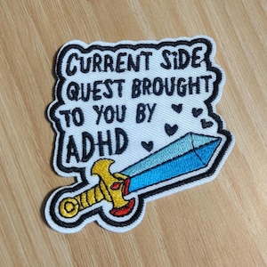 May include: Embroidered patch with the text "CURRENT SIDE QUEST BROUGHT TO YOU BY ADHD." The patch features a blue sword with a gold hilt and red accents, and small black heart shapes. The patch has a white background with a black border.