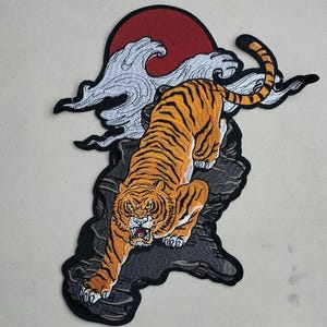 May include: An embroidered iron-on patch featuring a tiger with orange and black stripes, standing on a rocky outcrop with a white cloud and red sun behind it.