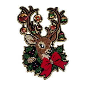 May include: A decorative iron-on patch featuring a brown reindeer head with large antlers adorned with hanging sequined ornaments and bells. The reindeer is framed by green holly leaves, red berries, and a large red bow at the base.