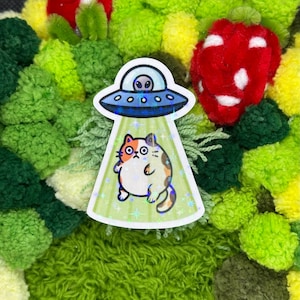 May include: A sticker featuring a cartoon cat being beamed up by a blue UFO. The cat is orange, white, and brown. The UFO has an alien inside. The sticker is set against a background of green and red pom-poms.