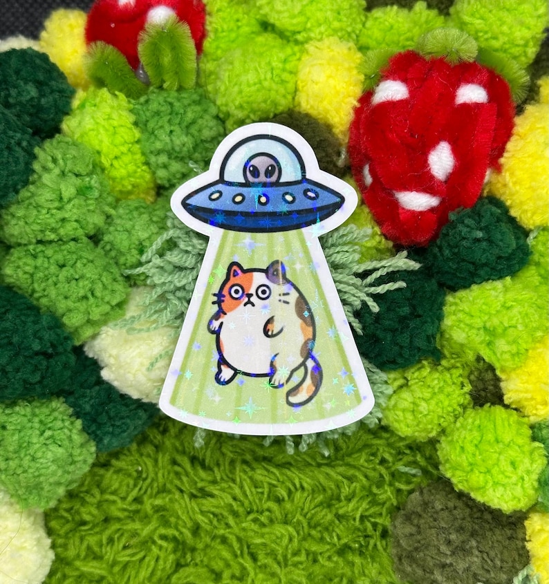 May include: A sticker featuring a cartoon cat being beamed up by a blue UFO. The cat is orange, white, and brown. The UFO has an alien inside. The sticker is set against a background of green and red pom-poms.