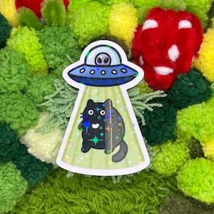 May include: A sticker featuring a black cat being beamed up by a UFO. The UFO is blue with a silver alien inside. The cat is black with white eyes and a tail. The background is a light green with stars.