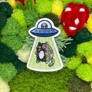 May include: A sticker featuring a cartoon cat being beamed up by a UFO. The UFO is blue with an alien inside. The cat is black, orange, and white. The background is a mix of green, yellow, and red pom-poms.