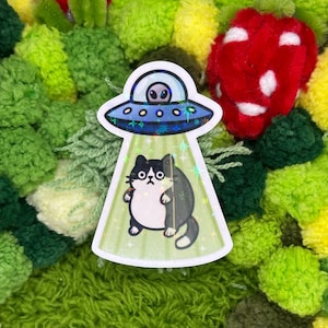 May include: A sticker featuring a black and white cat being abducted by a UFO. The UFO is silver and blue, with an alien inside. The background is a mix of green, yellow, and red pom-poms.