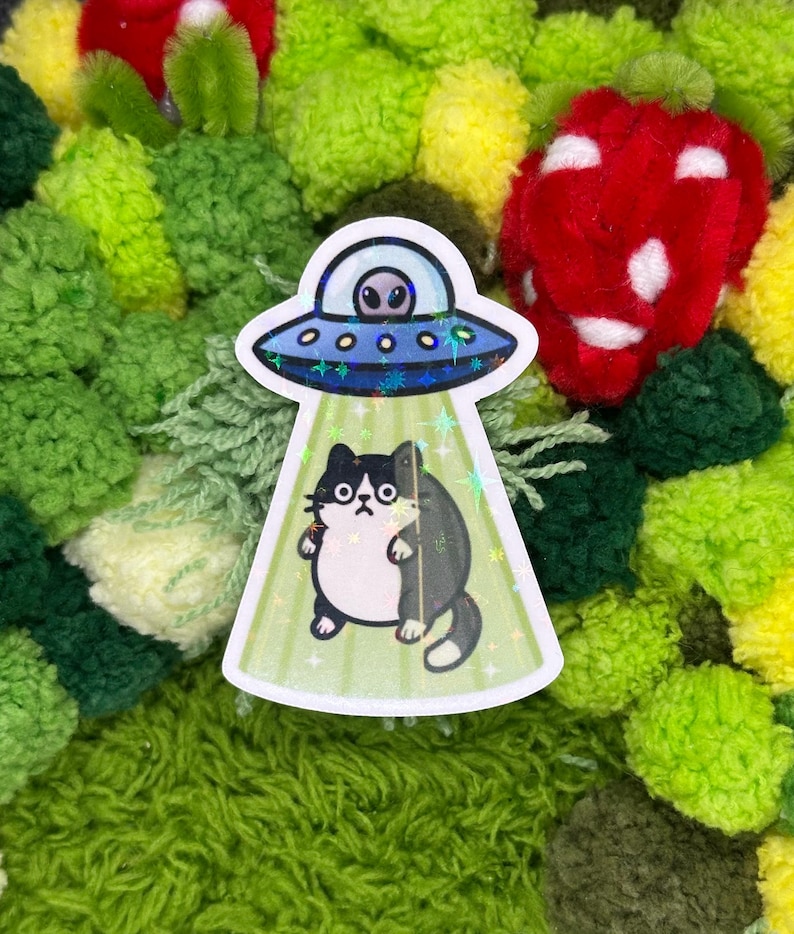 May include: A sticker featuring a black and white cat being abducted by a UFO. The UFO is silver and blue, with an alien inside. The background is a mix of green, yellow, and red pom-poms.