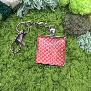 May include: A silver keychain with a square pendant featuring a red and pink snakeskin pattern. The keychain has a lobster clasp and a chain link. The background is a textured green fabric.