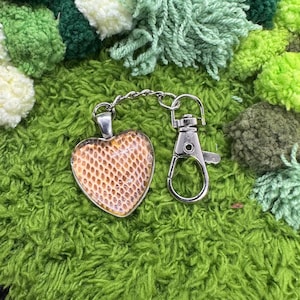 May include: A heart-shaped keychain pendant with a reptile skin pattern, connected to a silver-colored clasp. The pendant is set against a green, textured background, with various shades of green pom-poms.