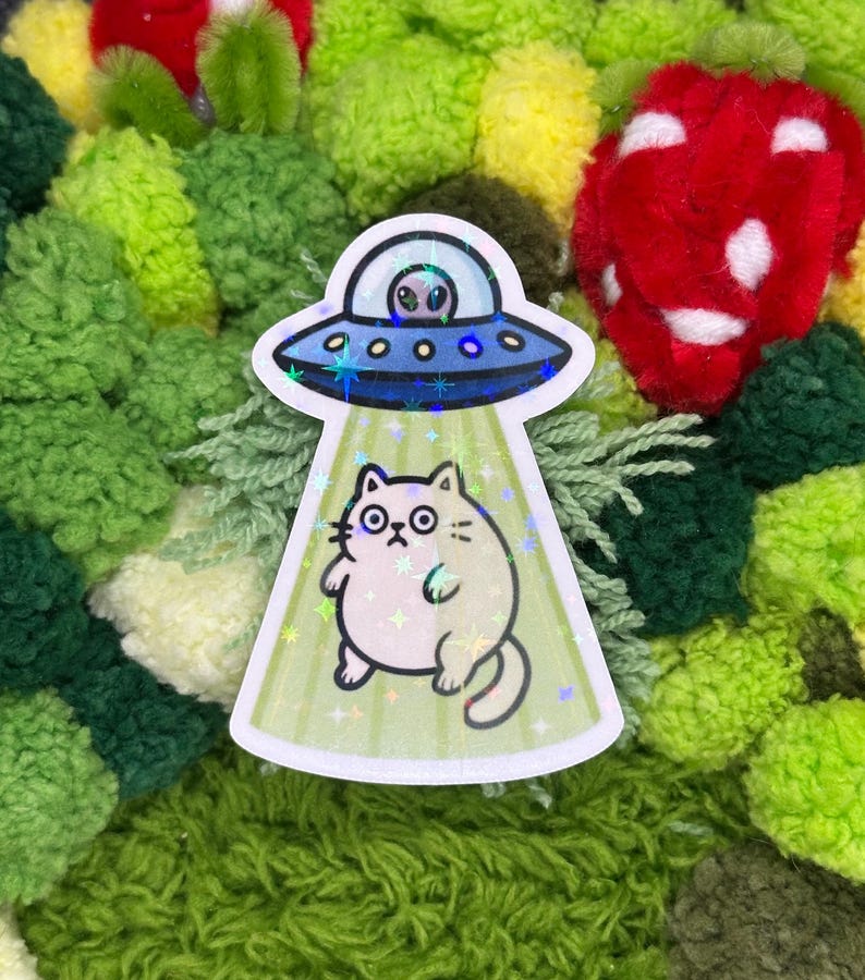 May include: A sticker featuring a cartoon cat being abducted by a UFO. The UFO is blue with a silver trim and an alien inside. The cat is light beige with large eyes and is being beamed up by a green light.