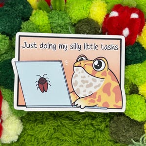 May include: A sticker featuring a cartoon frog working on a laptop. The frog is yellow and orange with large eyes. The laptop screen displays a bug. The text on the sticker reads, "Just doing my silly little tasks."