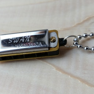 May include: A miniature harmonica pendant with the words "SWAN HARMONICA" engraved on the silver-coloured cover. The harmonica has a gold-coloured body and is attached to a silver ball chain necklace. The background is a light wood surface.
