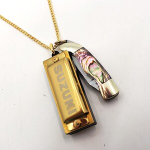 Può includere: Una collana con catena dorata con un ciondolo a forma di armonica dorata inciso con la parola "SUZUKI" e un piccolo ciondolo a forma di coltellino multi-colore.