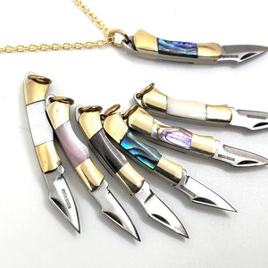 May include: A set of six small, decorative pocket knives with gold accents and different colored inlays. The knives are arranged in a fan shape and have a chain attached to one of them.
