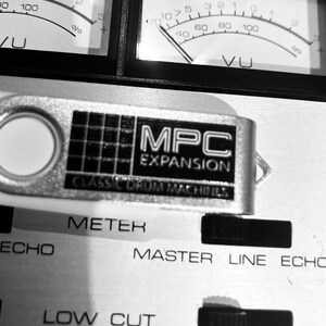 May include: A silver USB flash drive labeled MPC EXPANSION CLASSIC DRUM MACHINES resting on a metallic audio equipment panel featuring VU meters, METER, ECHO, MASTER LINE ECHO, and LOW CUT text labels in a black and white high contrast view.