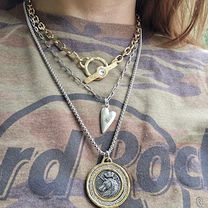May include: A layered necklace set featuring a gold-tone toggle chain with a clear stone, a silver-tone chain with a heart pendant displaying a small star, and a longer silver-tone chain with a circular pendant showing a unicorn and crown.