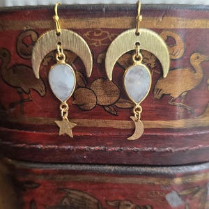 May include: A pair of gold-colored earrings featuring a crescent moon design at the top. Each earring has a teardrop-shaped, light-colored stone pendant, with a small star and moon charm dangling below. The earrings are displayed on a red and gold decorative surface.