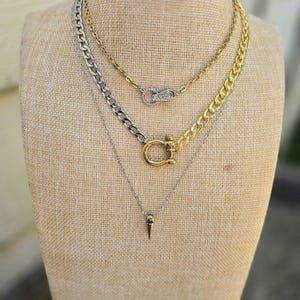 May include: A layered necklace set featuring three distinct chains. The top chain is gold-toned with a crystal-encrusted infinity clasp. The middle chain is a mixed-metal curb link with a gold-toned shackle clasp. The bottom chain is silver-toned with a small spike pendant.