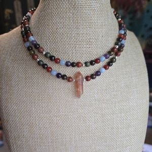 May include: A layered necklace with a variety of gemstone beads, including red, blue, green, and brown. The necklace has a pendant made of a light brown, pointed crystal.