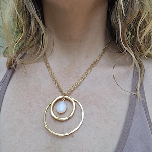 May include: Gold-toned necklace featuring three circular pendants. The largest pendant is a hammered-texture ring, followed by a smaller ring, and a teardrop-shaped pendant with a white stone. The necklace hangs on a delicate chain.