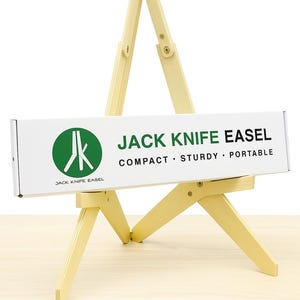 Könnte beinhalten: Hellfarbenes Jack Knife Easel aus Holz, kompakt, robust und tragbar. Das Staffelei hat eine weiße Box mit dem Produktnamen und einem grünen kreisförmigen Logo. Für Künstler konzipiert.