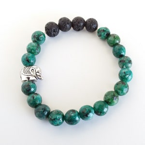 Puede incluir: Un brazalete de cuentas verdes y negras con un dije de elefante plateado. El brazalete está hecho con cuentas de lava y jaspe verde.