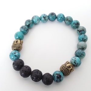 Puede incluir: Un brazalete de cuentas turquesa y negras con dos dijes de cabeza de Buda dorados. El brazalete está hecho con cuentas de lava y turquesa.