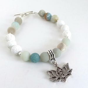 May include: A beaded bracelet with white lava beads, green and grey agate beads, and a silver charm of a lotus flower with an Om symbol.