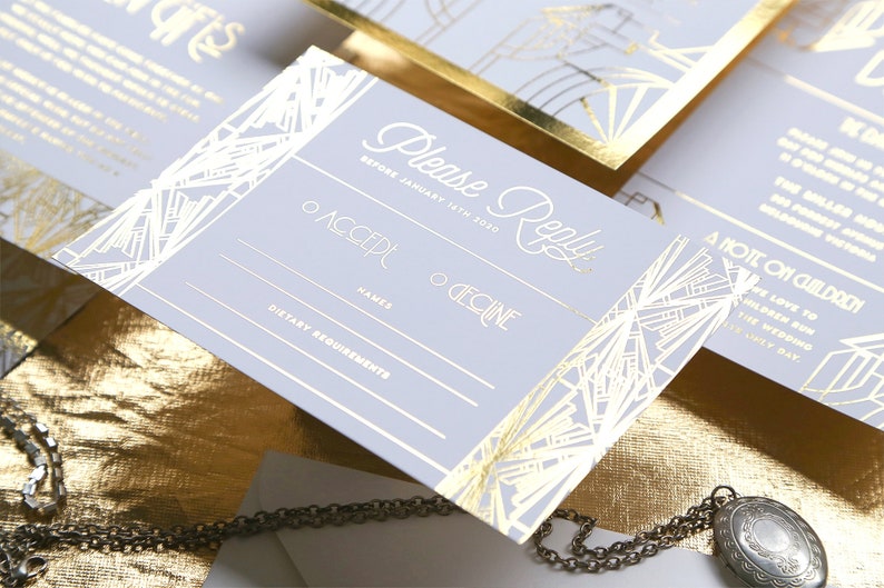 May include: Art Deco style wedding invitation set. The invitation and response card feature gold foil detailing on a pale grey background. The invitation includes the words "Please Reply" and RSVP information.