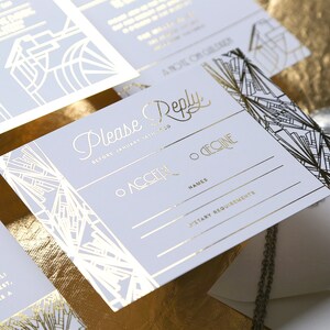 May include: A set of wedding stationery with a vintage art deco theme. The invitations and reply cards have gold foil detailing on a pale grey background. The cards feature geometric patterns and include the words "Please Reply".