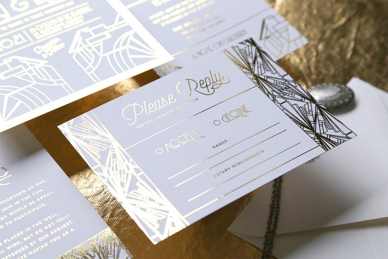 May include: A set of wedding stationery with a vintage art deco theme. The invitations and reply cards have gold foil detailing on a pale grey background. The cards feature geometric patterns and include the words "Please Reply".