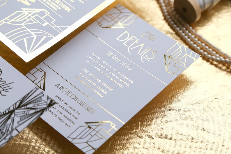 May include: Light grey wedding stationery with gold foil art deco detailing. The invitation text is in gold, including the date, time, and venue. The stationery is displayed on a gold textured surface.
