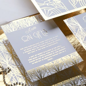 May include: A set of wedding stationery with a light grey and gold art deco design. The cards include the text "A Note on Gifts" and "Please RSVP". A gold ribbon and a decorative seal are also visible. The stationery is on a gold surface.