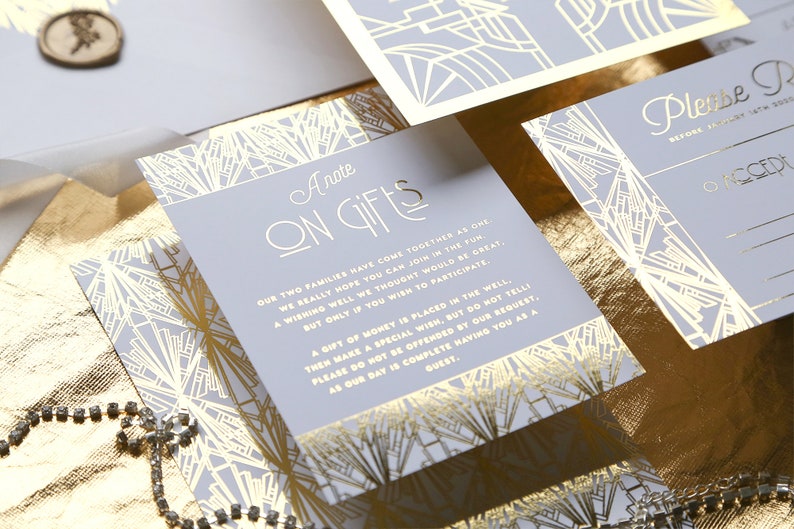 May include: A set of wedding stationery with a light grey and gold art deco design. The cards include the text "A Note on Gifts" and "Please RSVP". A gold ribbon and a decorative seal are also visible. The stationery is on a gold surface.