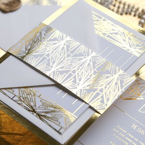 May include: A stylish wedding invitation suite with a gold art deco design. The invitation has a light grey background with gold foil accents and geometric patterns. The details are printed in gold, and the suite includes envelopes and inserts.