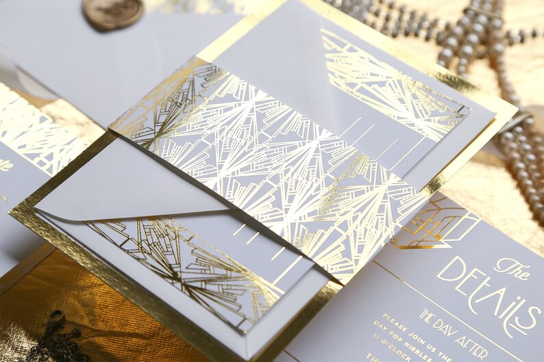 May include: A stylish wedding invitation suite with a gold art deco design. The invitation has a light grey background with gold foil accents and geometric patterns. The details are printed in gold, and the suite includes envelopes and inserts.