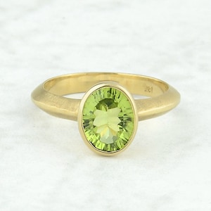 May include: A gold ring featuring a large, oval-cut, light green gemstone. The ring has a brushed gold band and a bezel setting that holds the gemstone securely. The ring is a piece of jewelry.