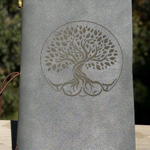 May include: A gray journal with a tree of life design embossed on the cover. The journal has a textured surface and a brown leather tie closure. The journal is held by a black metal stand.
