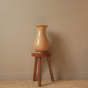 Peut inclure: Un vase en céramique brun clair posé sur un tabouret en bois à trois pieds. Le tabouret a un siège en cuir brun avec des clous en laiton. Le tabouret et le vase sont sur un tapis tissé brun clair.
