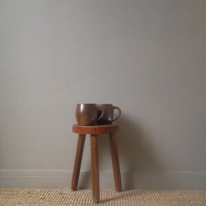 Peut inclure: Deux tasses en céramique marron sont posées sur un tabouret en bois à trois pieds. Le tabouret a un dessus en cuir marron et se trouve sur un tapis tissé.