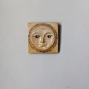 May include: A ceramic tile featuring a raised design of a smiling face with eyes and a mouth. The face is encircled by a ring of small brown dots.