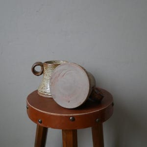 May include: Two ceramic mugs with a brown and white glaze, sitting on a brown leather stool with metal studs.
