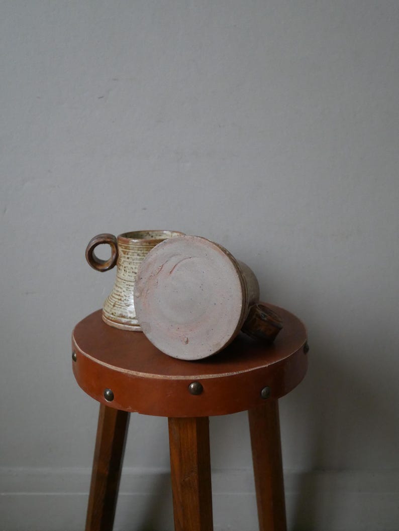 May include: Two ceramic mugs with a brown and white glaze, sitting on a brown leather stool with metal studs.